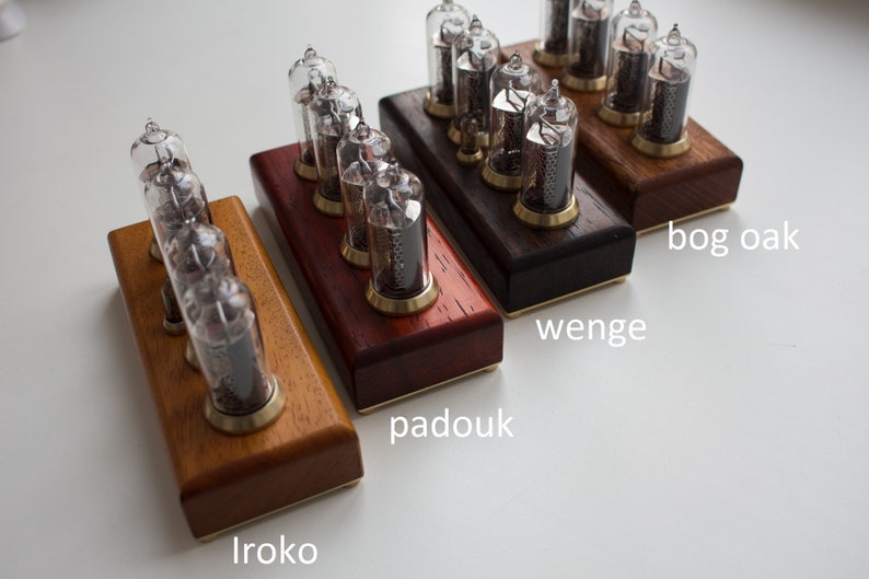 May include: Four wooden bases for a Nixie tube clock, each with a different wood type: Iroko, Padouk, Wenge, and Bog Oak. The bases are arranged in a row from left to right, with the Iroko base at the bottom and the Bog Oak base at the top.