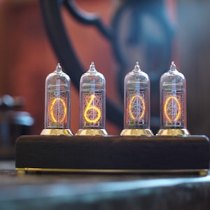 May include: A vintage-style clock with four glowing orange Nixie tubes displaying the time as 00:00. The clock is made of metal and has a dark wood base.