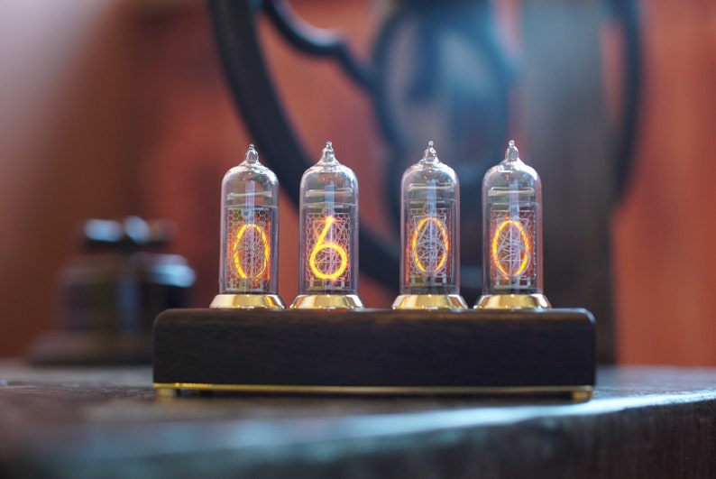 May include: A vintage-style clock with four glowing orange Nixie tubes displaying the time as 00:00. The clock is made of metal and has a dark wood base.