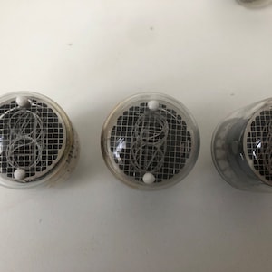 Puede incluir: Tres tubos Nixie de vidrio transparente con un patr&oacute;n de rejilla negra y puntos blancos en la parte superior. Los tubos se utilizan para mostrar n&uacute;meros.