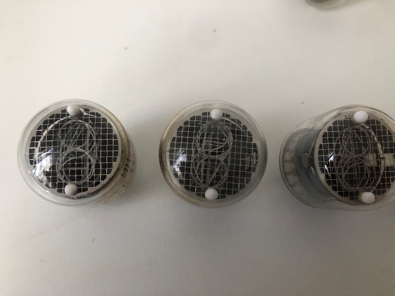 Puede incluir: Tres tubos Nixie de vidrio transparente con un patr&oacute;n de rejilla negra y puntos blancos en la parte superior. Los tubos se utilizan para mostrar n&uacute;meros.