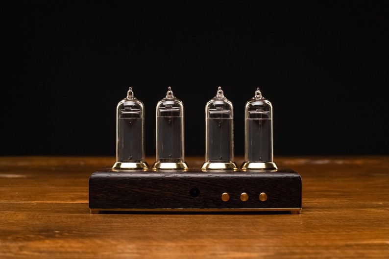 May include: A four-digit clock with clear glass tubes and a dark wood base. The tubes are lit up with a dark gray color, and the base has a gold trim.