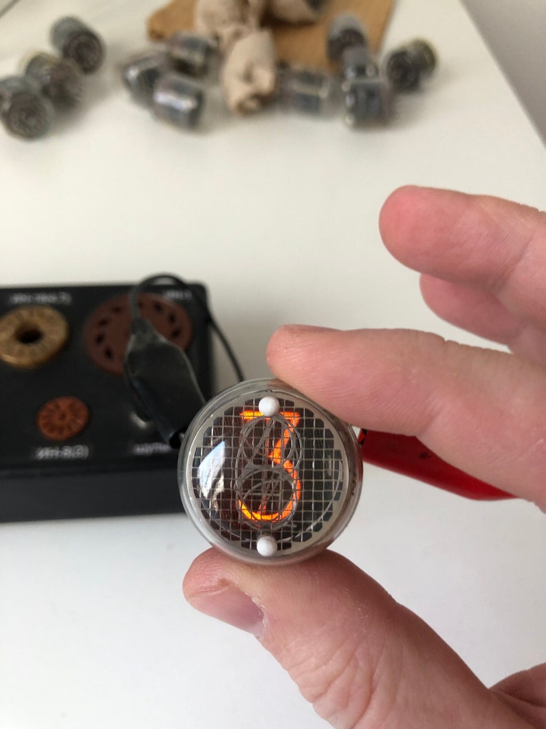 Puede incluir: Primer plano de un tubo Nixie redondo de color plateado que muestra el n&uacute;mero "13" en naranja. El tubo tiene dos puntos blancos en la parte superior e inferior.