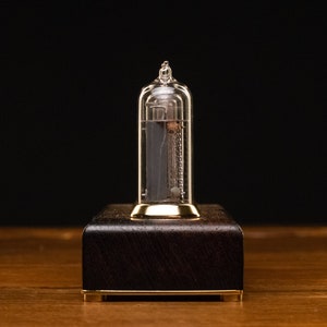 May include: A clear glass Nixie tube clock with a gold base and a dark wood frame. The tube is lit up and displaying the number 1.