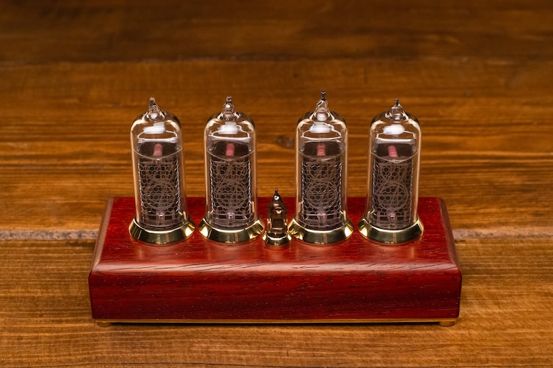 Puede incluir: Un reloj de estilo vintage con cuatro tubos Nixie de vidrio transparente que muestran la hora. El reloj est&aacute; montado sobre una base de madera roja oscura con detalles dorados.