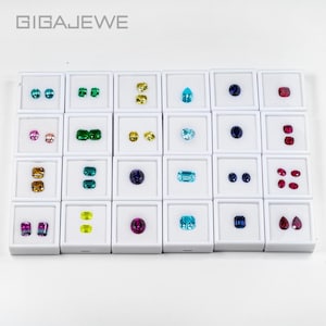 Può includere: Una collezione di pietre preziose in vari colori, tra cui blu, verde, giallo, rosa e rosso, esposte in singole scatole quadrate bianche. Il testo "GIGAEWE" è visibile in alto. Le pietre preziose sono di diverse forme e dimensioni.