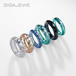May include: A selection of six gemstone rings in a range of colours: clear, gold, blue, teal, black, and green. The rings are displayed diagonally on a white background. The brand name "GIGAEWE" is visible at the top.