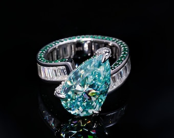 May include: A silver ring with a large pear-shaped blue gemstone surrounded by smaller green gemstones. The ring is set on a black background.