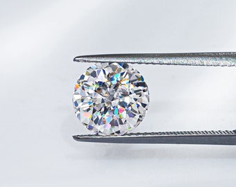 May include: A clear round diamond gemstone held by silver tweezers against a white background. The diamond has a rainbow effect.