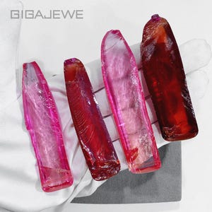 May include: Four elongated, translucent, raw gemstone specimens in varying shades of red and pink, ranging from light magenta to deep crimson. The stones have a smooth, polished appearance with visible internal fractures. The text GIGAJEWE is visible.