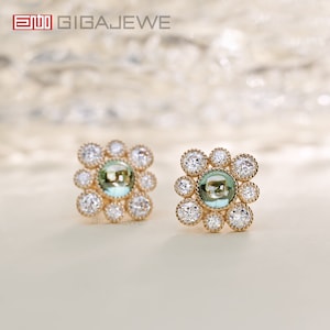 May include: A pair of gold earrings with a square design. Each earring features a round, light green gemstone surrounded by smaller, clear gemstones.