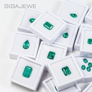 Peut inclure: Plusieurs pierres précieuses d'émeraude de différentes formes, dont des tailles poire et rectangulaires, présentées dans des boîtes blanches individuelles. Le texte "GIGAEWE" est visible. Les pierres sont d'un vert vif.