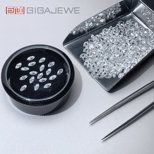 May include: A black round container with a clear lid filled with small, clear, marquise-shaped gemstones. A metal scoop filled with the same gemstones is next to the container. Two silver tweezers are also in the image.
