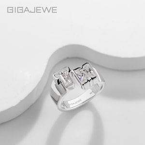 Puede incluir: Un anillo de tono plateado con dos letras abiertas estilo bloque, T y M, cada una incrustada con piedras claras de corte rectangular. La banda interior está grabada con el texto 925 y GIGAJEWE, indicando una composición de plata esterlina.
