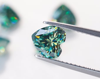 Op de afbeelding: Een hartvormige groene moissanite-edelsteen vastgehouden door een pincet. De edelsteen heeft een briljant-slijpsel en een stervormig patroon in het midden.
