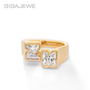 Può includere: Un anello d'oro con due pietre preziose quadrate e trasparenti. L'anello ha una fascia spessa e le pietre preziose sono incastonate in un design geometrico unico. Il marchio "GIGAEWE" si trova nell'angolo in alto a sinistra.