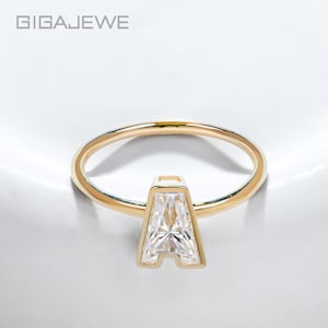 Può includere: Un anello d'oro con una gemma trasparente, taglio rettangolare, a forma di lettera "A". L'anello ha una fascia sottile e lucida. Il nome del marchio "GIGAEWE" è visibile in alto a sinistra.