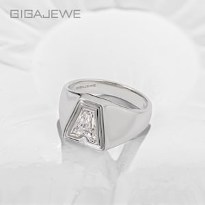 Puede incluir: Un anillo de sello en tono plateado con un engaste prominente en forma de letra A. El centro de la letra A está incrustado con una piedra transparente facetada. El interior de la banda tiene grabado el nombre de la marca GIGAJEWE, visible arriba.