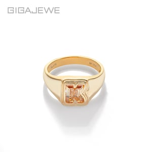 Puede incluir: Anillo dorado con una gema rectangular de color naranja claro en forma de la letra "R". El anillo tiene un acabado pulido y está sobre un fondo blanco. El nombre de la marca "GIGAEWE" es visible en la esquina superior izquierda.