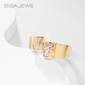 May include: A gold-toned ring featuring two offset, rectangular-cut clear stones set within a smooth, polished metal band. The brand name GIGAJEWE is printed in the upper left corner of the frame. The design showcases a modern, open-ended structure.
