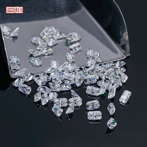Puede incluir: Una colección de gemas claras y facetadas, incluyendo formas rectangulares y de diamante, se dispersan sobre una cuchara plateada y una superficie oscura y reflectante. Las gemas brillan, reflejando la luz. El logotipo "GIGAJEWE" es visible en la esquina superior izquierda.