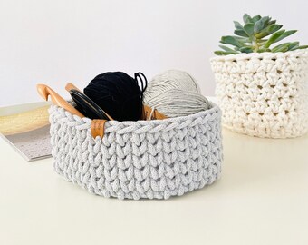 Könnte beinhalten: Ein grauer Häkelkorb mit einem Holzgriff, gefüllt mit Wollknäueln und Stricknadeln. Der Korb steht auf einem weißen Tisch neben einem weißen Häkelkorb mit einer Sukkulentenpflanze.