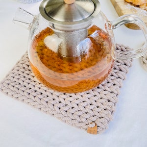 May include: A glass teapot with a stainless steel lid and a wooden knob. The teapot is filled with amber-coloured tea and is sitting on a beige crocheted coaster.