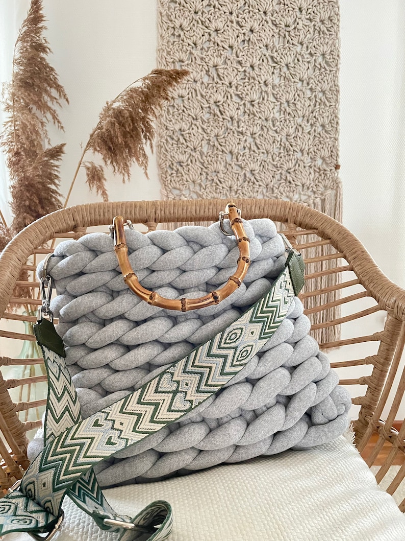 May include: A gray knitted tote bag with a bamboo handle and a green and white patterned strap. The bag is sitting on a wicker chair.