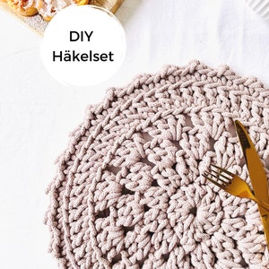 Könnte beinhalten: DIY Häkelset - Nahaufnahme eines gehäkelten Platzsets in hellrosa. Das Platzset ist rund und hat ein filigranes, kompliziertes Design. Eine goldene Gabel und ein Messer liegen auf dem Platzset.