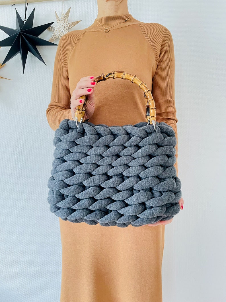 May include: A gray knitted tote bag with a bamboo handle. The bag is made of thick, chunky yarn and has a woven texture.