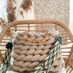 May include: A beige knitted tote bag with a bamboo handle and a green and white patterned strap. The bag is sitting on a wicker chair.