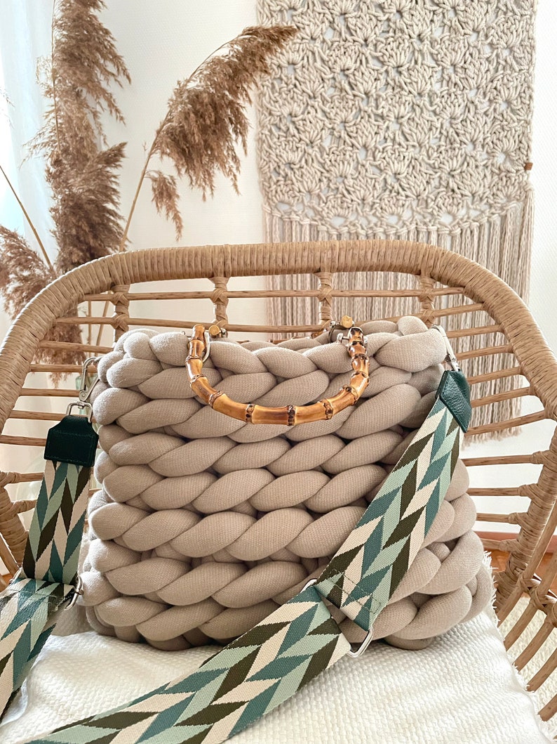 May include: A beige knitted tote bag with a bamboo handle and a green and white patterned strap. The bag is sitting on a wicker chair.