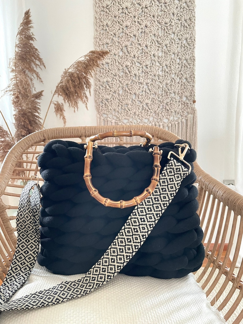 May include: Black knitted tote bag with a bamboo handle and a black and white patterned strap. The bag is sitting on a wicker chair.