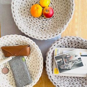 K&ouml;nnte beinhalten: Drei runde, geflochtene K&ouml;rbe in Beige und Wei&szlig;. Der gr&ouml;&szlig;te Korb ist mit drei Orangen und einem roten Apfel gef&uuml;llt. Der mittlere Korb ist mit einer Sonnenbrille, einem Handy-Etui und einer kleinen grauen Tasche gef&uuml;llt. Der kleinste Korb ist mit einem Stapel B&uuml;cher und einem Magazin mit dem Wort "GU" auf dem Cover gef&uuml;llt.