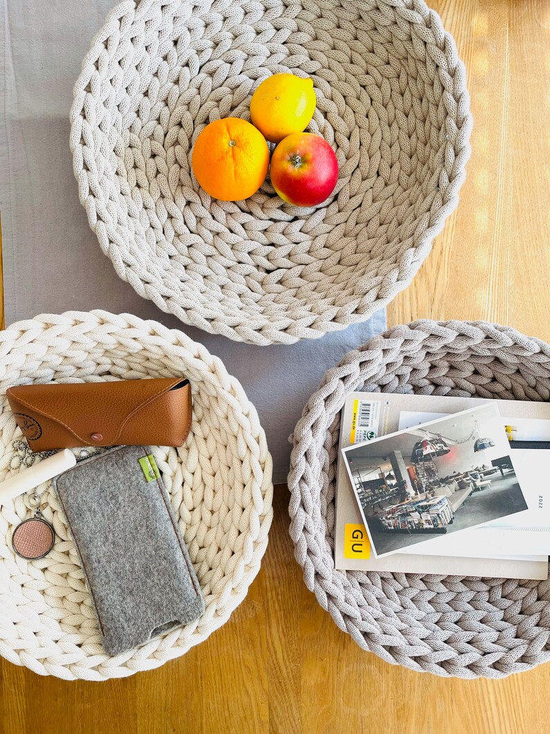 K&ouml;nnte beinhalten: Drei runde, geflochtene K&ouml;rbe in Beige und Wei&szlig;. Der gr&ouml;&szlig;te Korb ist mit drei Orangen und einem roten Apfel gef&uuml;llt. Der mittlere Korb ist mit einer Sonnenbrille, einem Handy-Etui und einer kleinen grauen Tasche gef&uuml;llt. Der kleinste Korb ist mit einem Stapel B&uuml;cher und einem Magazin mit dem Wort "GU" auf dem Cover gef&uuml;llt.