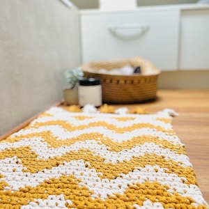 Puede incluir: Una alfombra de crochet blanca y mostaza con un patrón de chevron. La alfombra está sobre un suelo de madera en un baño.