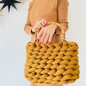 May include: A mustard yellow knitted tote bag with bamboo handles. The bag is made of thick, chunky yarn and has a woven texture.