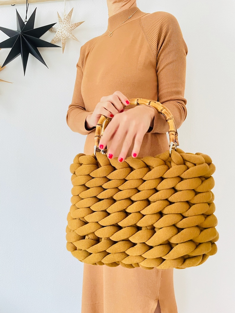 May include: A mustard yellow knitted tote bag with bamboo handles. The bag is made of thick, chunky yarn and has a woven texture.
