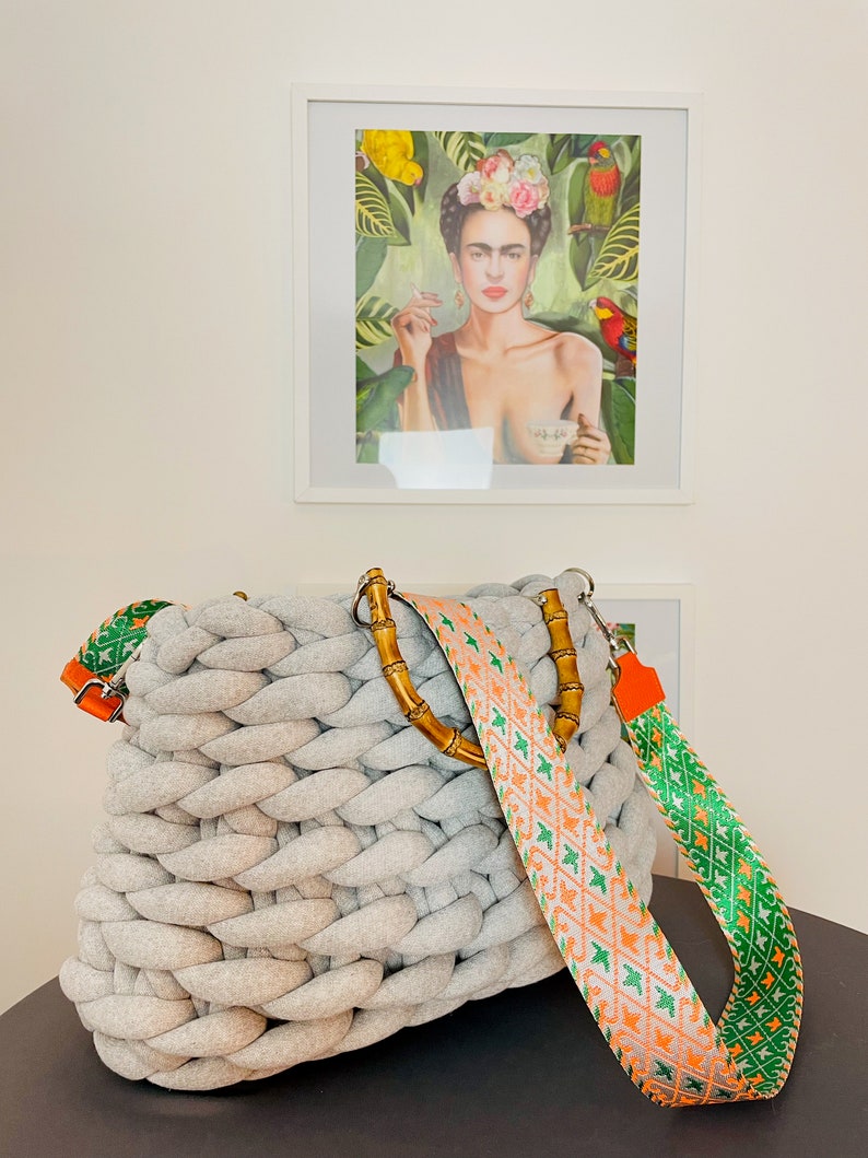 May include: A gray knitted tote bag with a bamboo handle and a colorful patterned strap. The strap is orange, green, and white with a geometric design.