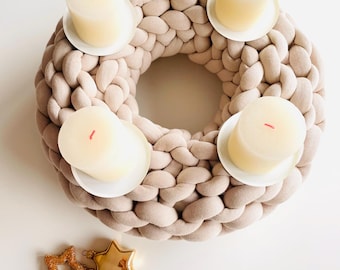 Puede incluir: Una corona de tela trenzada beige con cuatro velas color crema sobre platillos blancos. Hay adornos dorados en forma de estrella y bola en la parte inferior. La corona es una decoración festiva para la temporada navideña.