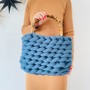 May include: A blue knitted tote bag with a bamboo handle. The bag is made of thick, chunky yarn and has a woven texture.