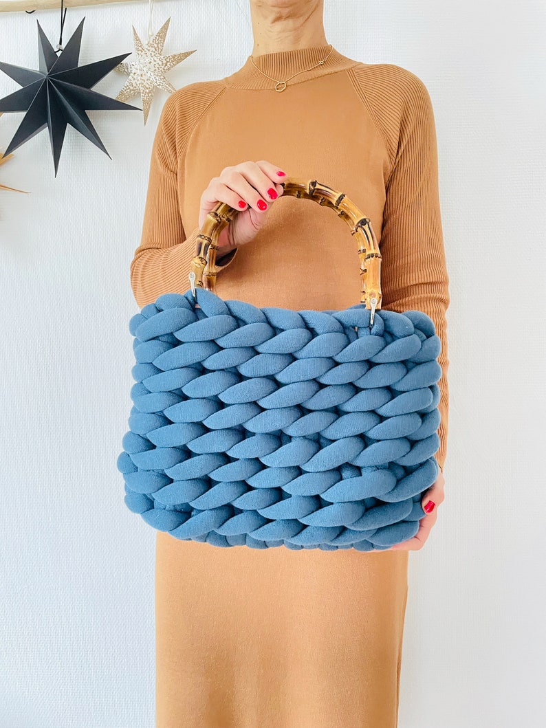May include: A blue knitted tote bag with a bamboo handle. The bag is made of thick, chunky yarn and has a woven texture.