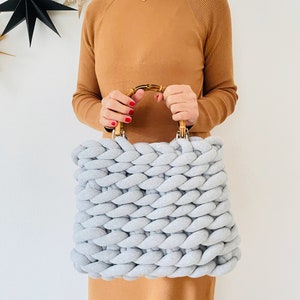 May include: A light gray knitted tote bag with a bamboo handle. The bag is made with thick, chunky yarn and has a square shape.