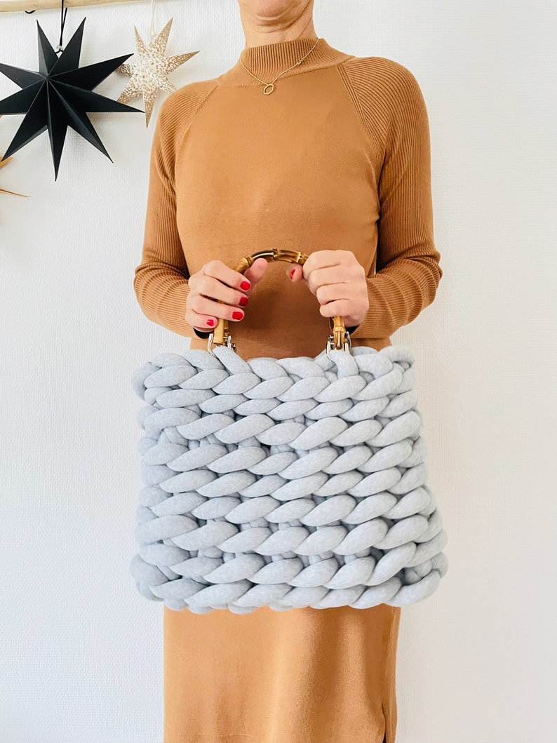 May include: A light gray knitted tote bag with a bamboo handle. The bag is made with thick, chunky yarn and has a square shape.