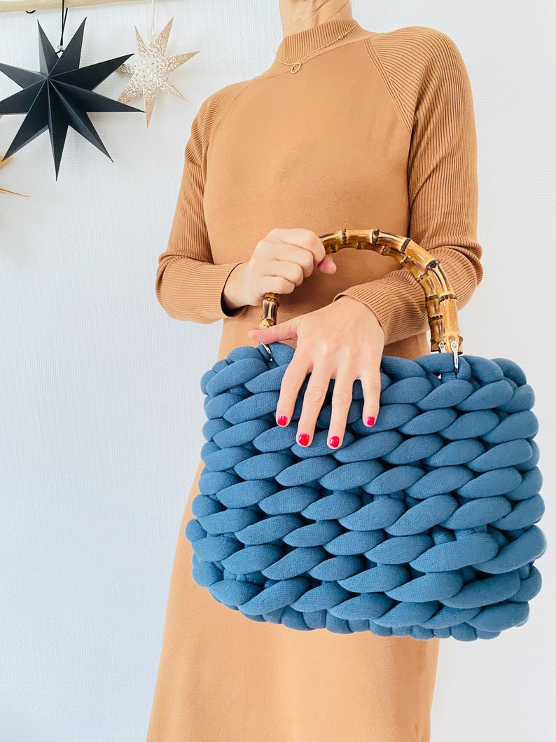 May include: A blue knitted tote bag with a bamboo handle. The bag is made of thick, chunky yarn and has a woven texture.