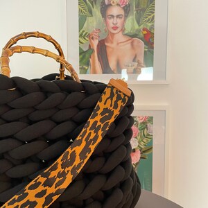 May include: Black knitted tote bag with a bamboo handle and a leopard print strap. The bag is sitting on a table in front of a framed print of Frida Kahlo.