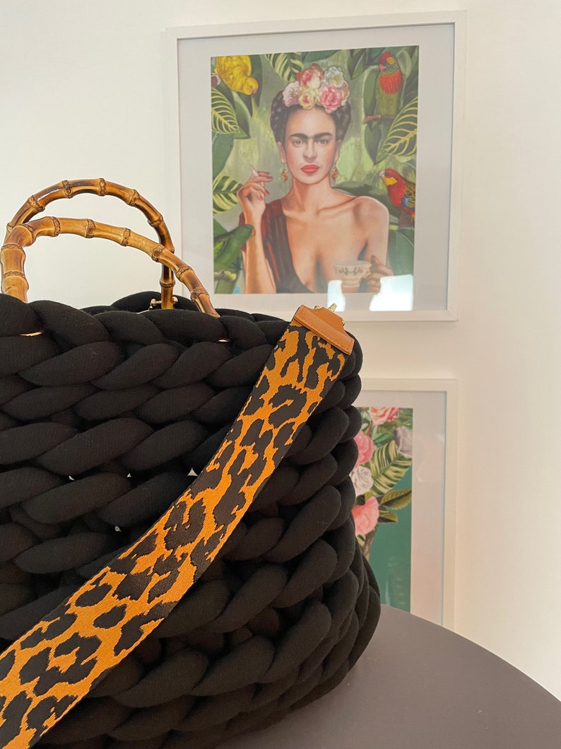 May include: Black knitted tote bag with a bamboo handle and a leopard print strap. The bag is sitting on a table in front of a framed print of Frida Kahlo.