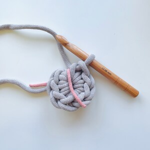 K&ouml;nnte beinhalten: Nahaufnahme eines grauen H&auml;kelkreises mit einem durchlaufenden rosa Garnfaden. Ein H&auml;kelnadel aus Holz mit dem Text "15.00 mm" ist im Bild zu sehen.