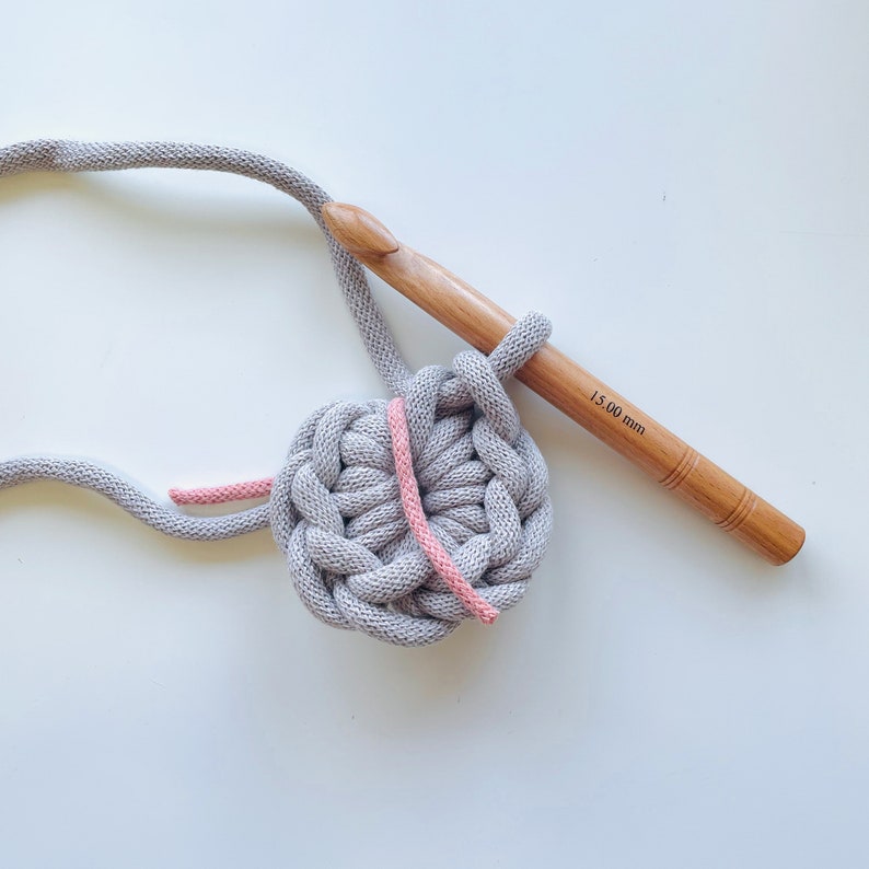 K&ouml;nnte beinhalten: Nahaufnahme eines grauen H&auml;kelkreises mit einem durchlaufenden rosa Garnfaden. Ein H&auml;kelnadel aus Holz mit dem Text "15.00 mm" ist im Bild zu sehen.