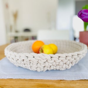 K&ouml;nnte beinhalten: Eine beige geh&auml;kelte Schale gef&uuml;llt mit Orangen und &Auml;pfeln steht auf einem hellblauen Platzdeckchen auf einem Holztisch.